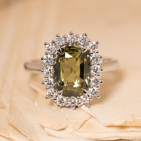 Antique-inspired green sapphire and diamond cluster engagement ring featuring a cushion-cut green sapphire surrounded by a halo of brilliant-cut diamonds in white gold.