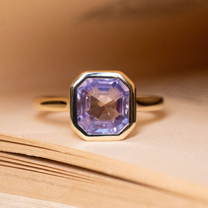 Antique-inspired lavender sapphire solitaire engagement ring featuring an emerald-cut lavender sapphire in a yellow gold rub-over setting.