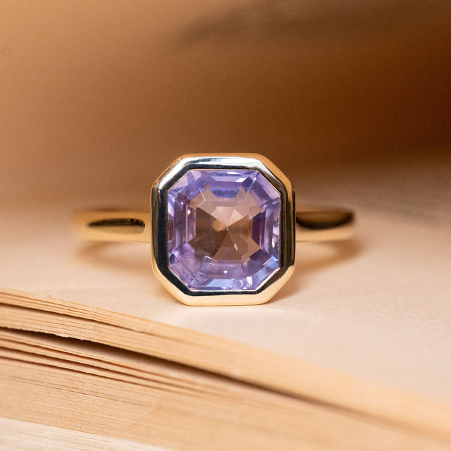 Antique-inspired lavender sapphire solitaire engagement ring featuring an emerald-cut lavender sapphire in a yellow gold rub-over setting.