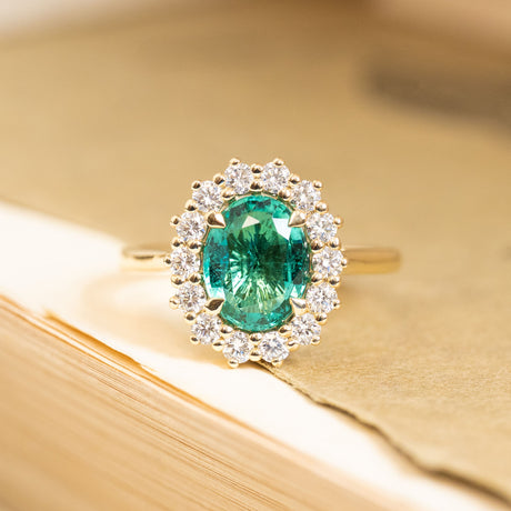 Antique-inspired oval-cut emerald and diamond cluster engagement ring featuring a vibrant green emerald surrounded by brilliant-cut diamonds in yellow gold.