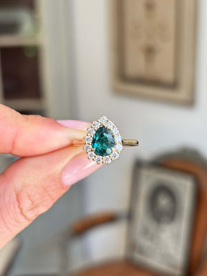 Antique-inspired pear-cut teal tourmaline and diamond cluster engagement ring with a pear-shaped teal tourmaline surrounded by brilliant-cut diamonds, held by hand.