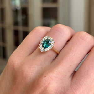 Antique-inspired pear-cut teal tourmaline and diamond cluster engagement ring featuring a pear-shaped teal tourmaline surrounded by brilliant-cut diamonds, displayed on a hand with a blurred background.