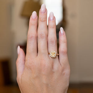 Antique inspired primrose-yellow sapphire and diamond cluster engagement ring worn on the hand, featuring an oval yellow sapphire surrounded by brilliant-cut diamonds in yellow gold.