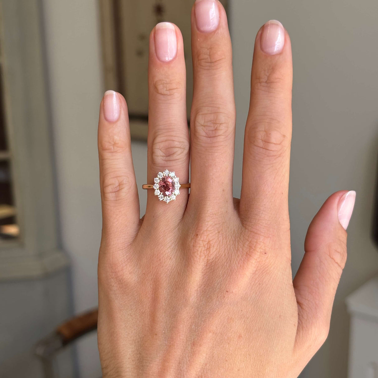 Antique-inspired rose-pink tourmaline and diamond cluster engagement ring featuring an oval rose-pink tourmaline encircled by brilliant-cut diamonds, displayed on a hand.