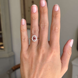 Antique-inspired rose-pink tourmaline and diamond cluster engagement ring featuring an oval rose-pink tourmaline encircled by brilliant-cut diamonds, displayed on a hand.