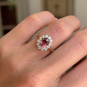 Antique-inspired rose-pink tourmaline and diamond cluster engagement ring with an oval rose-pink tourmaline centre and diamond halo, worn on a hand.