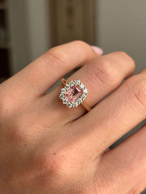 Antique-inspired sunset pink sapphire and diamond cluster engagement ring featuring an octagon-cut sunset pink sapphire encircled by brilliant-cut diamonds, worn on a hand.