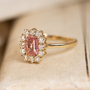 Antique-inspired sunset pink sapphire and diamond cluster engagement ring featuring an emerald-cut pink sapphire surrounded by brilliant-cut diamonds in yellow gold.