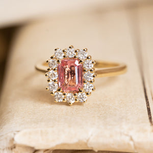 Sunset pink sapphire engagement ring set with an emerald-cut pink sapphire framed by a sparkling diamond cluster in yellow gold.