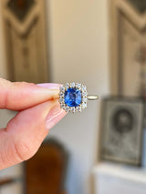 Antique-inspired violet-blue cushion cut Sri Lankan sapphire and diamond cluster engagement ring in yellow gold, held between fingers against a softly blurred vintage interior background, showcasing the vivid sapphire and sparkling diamond halo.