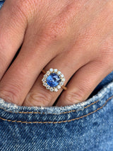 Antique-inspired violet-blue cushion cut Sri Lankan sapphire and diamond cluster engagement ring in yellow gold photographed on hand against blue denim, showcasing the vibrant sapphire centre stone and sparkling diamond cluster.