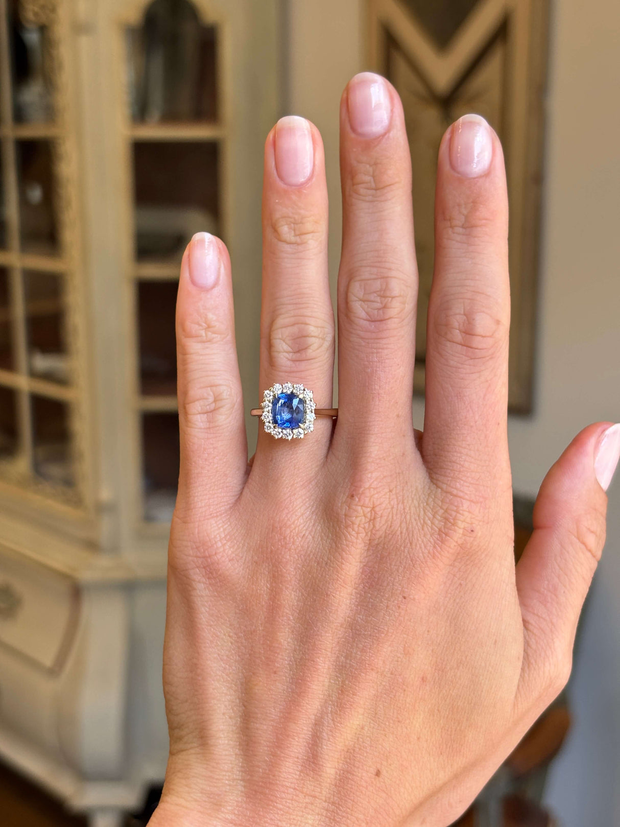 Antique-inspired violet-blue cushion cut Sri Lankan sapphire and diamond cluster engagement ring in yellow gold worn on hand, highlighting the vibrant blue sapphire centre stone and elegant diamond cluster setting against a vintage-inspired backdrop.