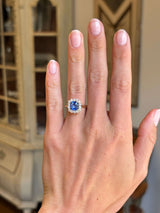 Antique-inspired violet-blue cushion cut Sri Lankan sapphire and diamond cluster engagement ring in yellow gold worn on hand, highlighting the vibrant blue sapphire centre stone and elegant diamond cluster setting against a vintage-inspired backdrop.