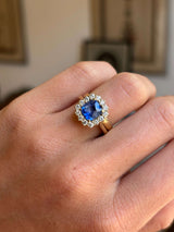 Antique-inspired violet-blue cushion cut Sri Lankan sapphire and diamond cluster engagement ring in yellow gold worn on hand with a gold wedding band, against a softly lit interior setting. 