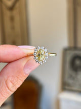 Antique-inspired yellow sapphire and diamond cluster engagement ring featuring an oval yellow sapphire surrounded by a halo of brilliant-cut diamonds, held by hand.