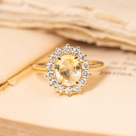 Antique-inspired pastel yellow sapphire and diamond cluster engagement ring featuring an oval-cut yellow sapphire surrounded by brilliant-cut diamonds in yellow gold.