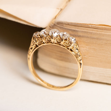 Antique Victorian five-stone ring featuring graduating old-mine cut diamonds with intricate hand-carved gold scrollwork shoulders.