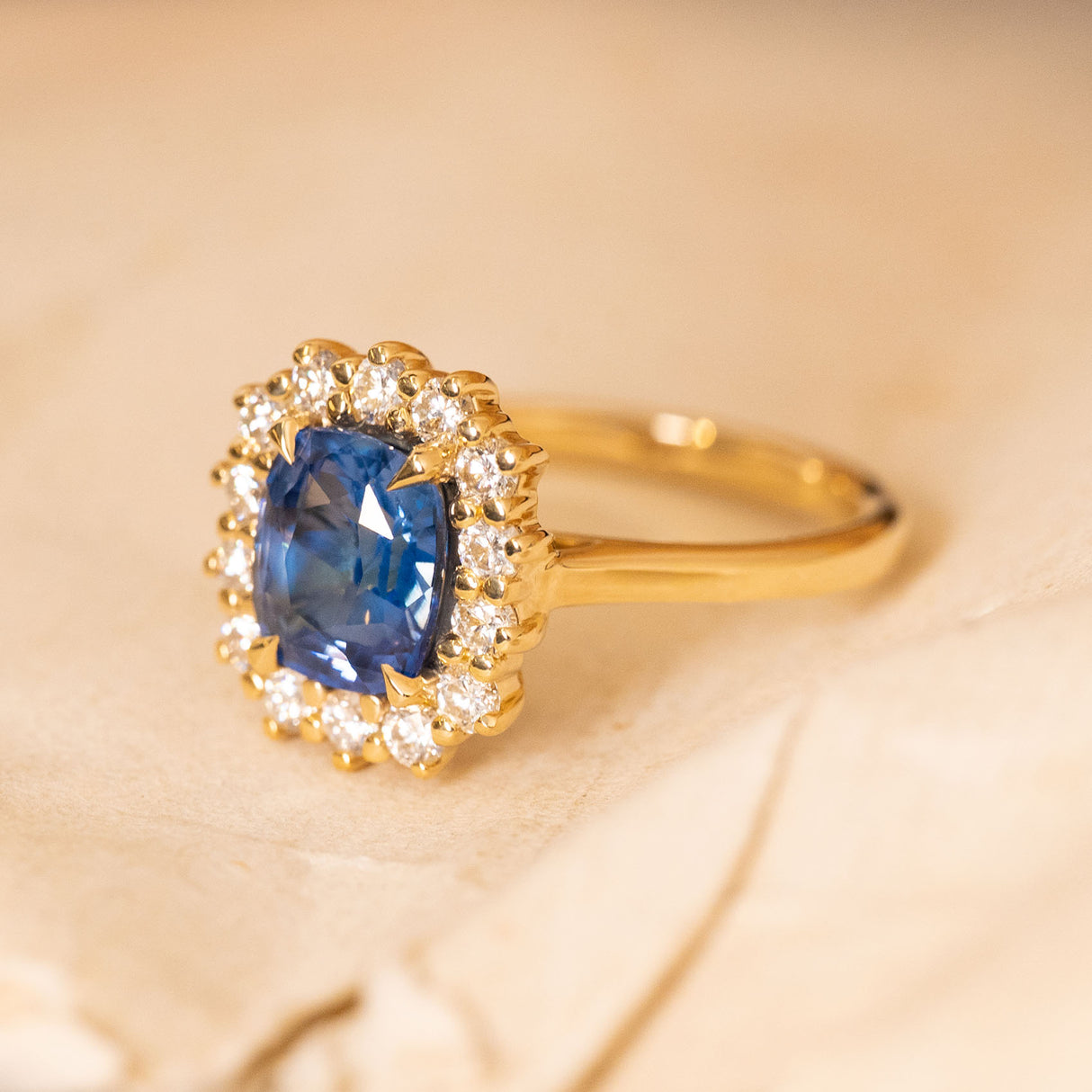 Antique-inspired sapphire and diamond cluster engagement ring with a cushion-cut blue sapphire framed by a halo of brilliant-cut diamonds.