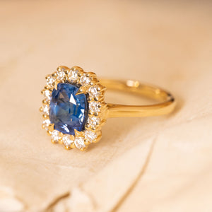 Antique-inspired sapphire and diamond cluster engagement ring with a cushion-cut blue sapphire framed by a halo of brilliant-cut diamonds.