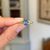 Blue sapphire and diamond engagement ring featuring an oval blue sapphire centre stone with diamond-set trefoil shoulders in the form of laurel leaves, held by hand.