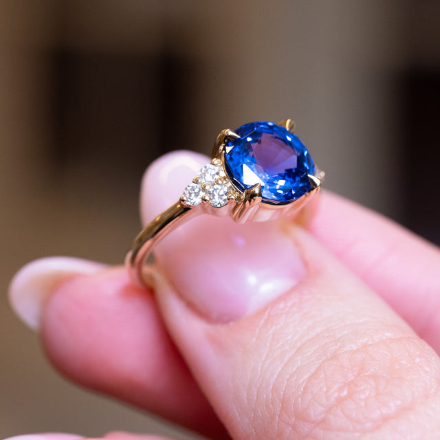 Round blue sapphire and diamond cluster tre-foil engagement ring with vivid blue sapphire and diamond side stones.