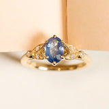 Blue sapphire and diamond engagement ring featuring an oval-cut sapphire set between diamond-set leaf motifs in yellow gold.