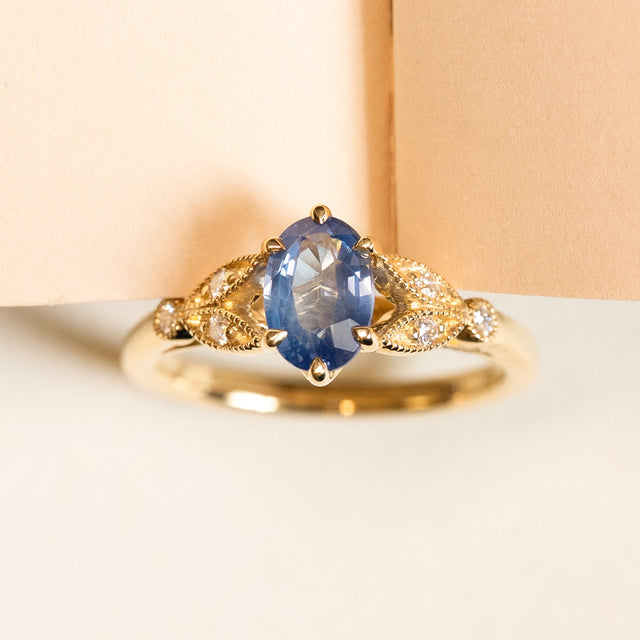 Blue sapphire and diamond engagement ring featuring an oval-cut sapphire set between diamond-set leaf motifs in yellow gold.
