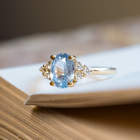 Pale blue sapphire and diamond cluster tre-foil engagement ring displayed on open book pages.