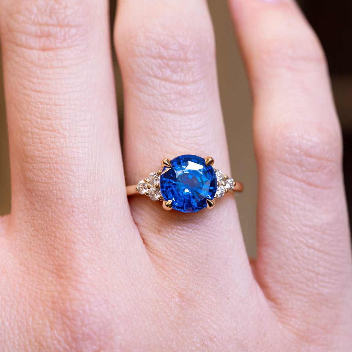 Round blue sapphire and diamond cluster tre-foil engagement ring with diamond-set shoulders worn on finger.