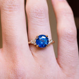 Round blue sapphire and diamond cluster tre-foil engagement ring with diamond-set shoulders worn on finger.