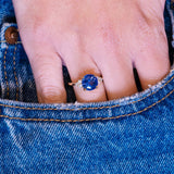 Round blue sapphire and diamond cluster tre-foil engagement ring worn on hand with denim pocket styling.