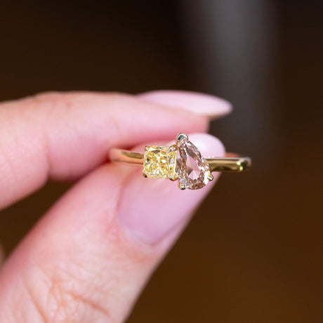 Brown and yellow diamond Toi et Moi engagement ring held between fingers with gold band.