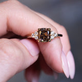 Chocolate brown diamond cluster tre-foil engagement ring held between fingers with gold band.