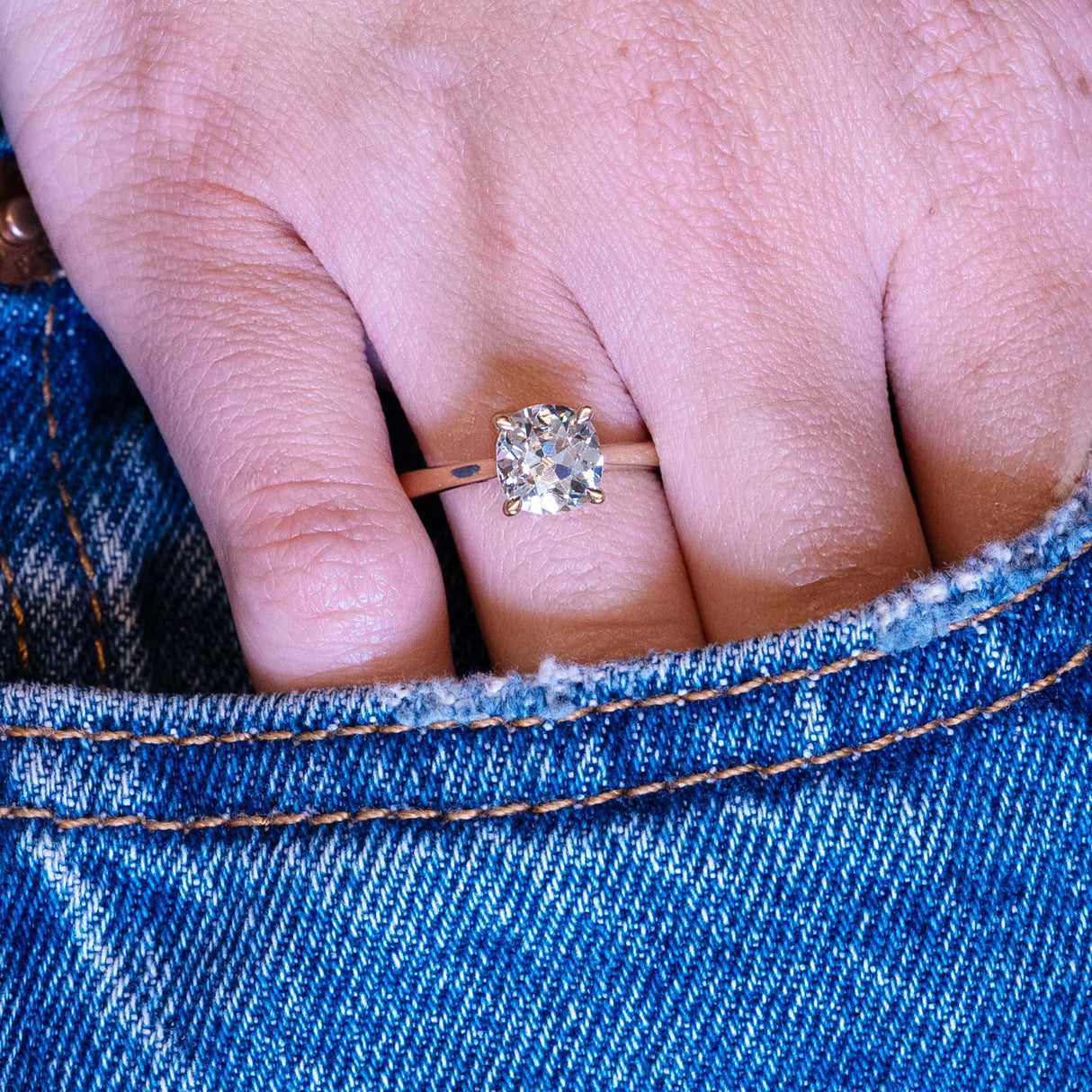 Cushion old-mine cut diamond solitaire engagement ring worn on hand with denim pocket styling.