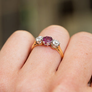 Gold ring with a central ruby and two diamonds on a finger against a blurred background.