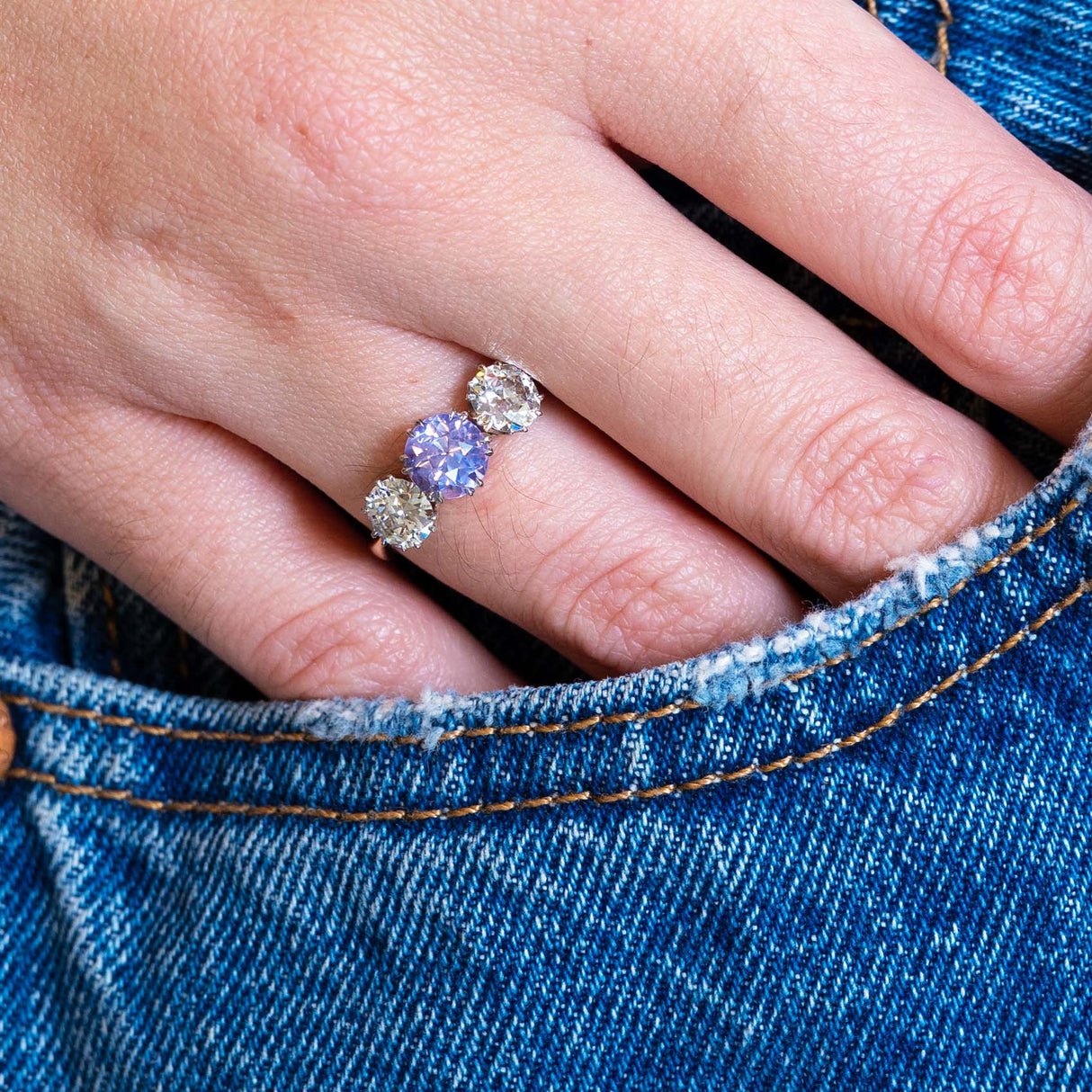 Close-up of a hand wearing a three-stone ring with a sapphire, set against a blue denim background.