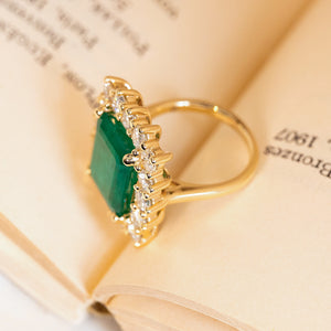Emerald and diamond cluster cocktail ring with a substantial emerald-cut emerald and dazzling diamond surround set in yellow gold.