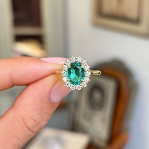 Close-up of an antique-inspired yellow gold emerald and diamond cluster ring, showcasing a vibrant oval-cut emerald framed by sparkling diamonds against a warm interior setting.