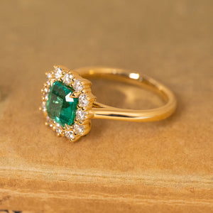 Emerald and diamond cluster engagement ring with an emerald-cut emerald encircled by brilliant-cut diamonds in a yellow gold setting.
