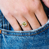 Green sapphire and diamond cluster engagement ring worn with hand resting in denim pocket.