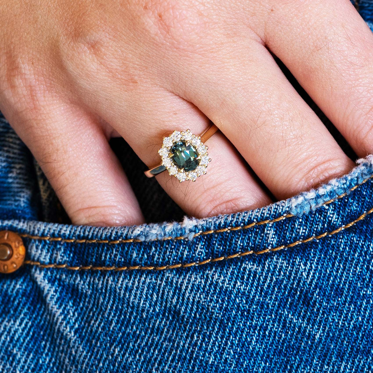 Close-up of a hand wearing a ring with a green sapphire, set against a denim background.