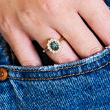 Close-up of a hand wearing a ring with a green sapphire, set against a denim background.