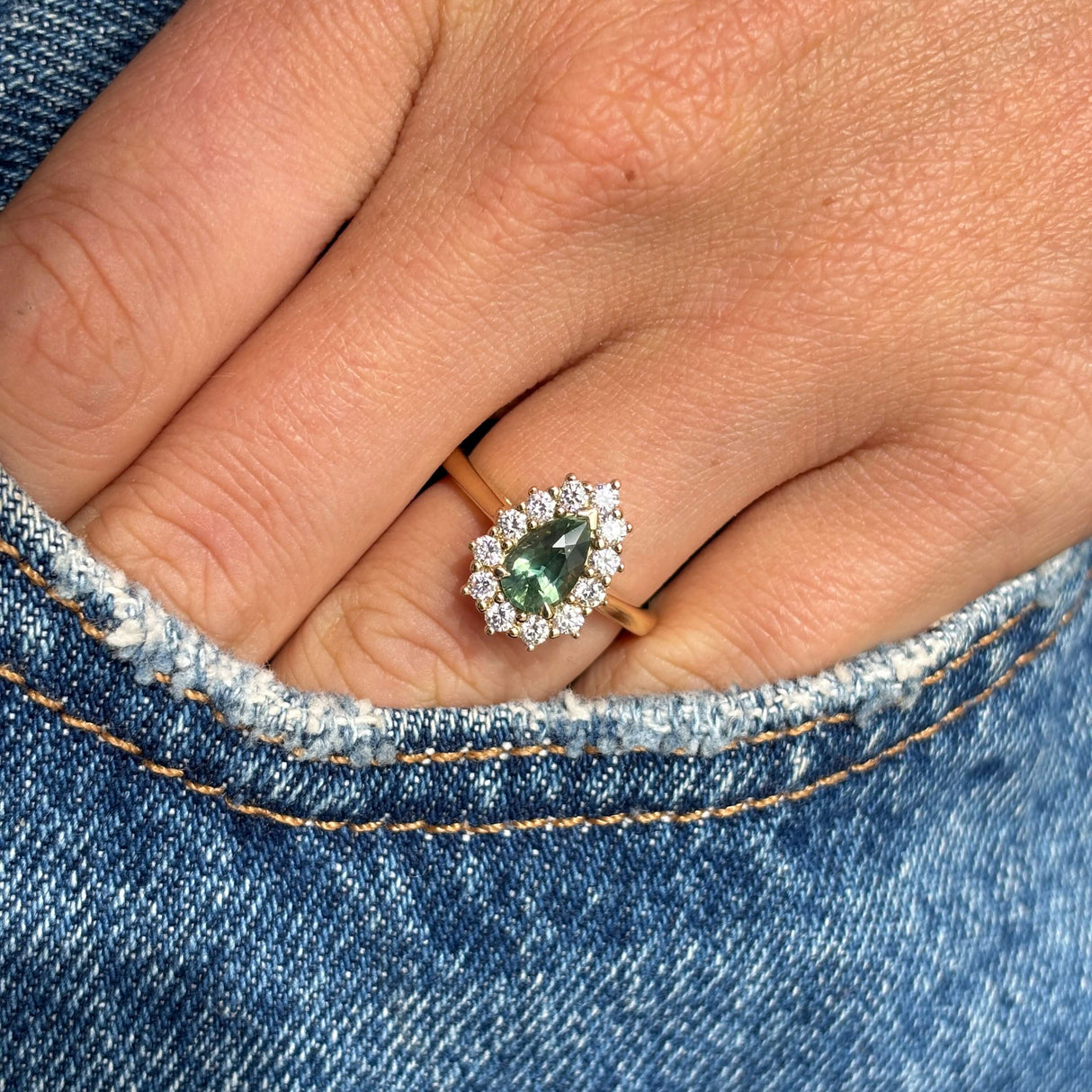 Antique-inspired yellow gold diamond cluster ring with a pear-cut green sapphire centre stone, styled on hand against blue denim to highlight colour, sparkle and scale.