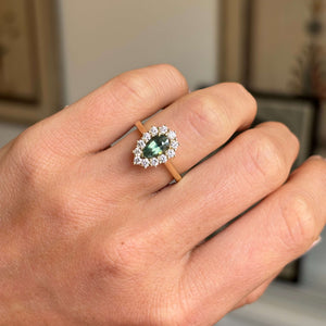 Close-up of a yellow gold sapphire cluster ring featuring a pear-cut green sapphire framed by bright diamonds, photographed against a warm interior setting.