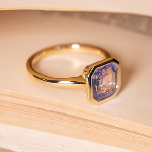 Antique-inspired lavender sapphire solitaire ring showcasing an emerald-cut lavender sapphire in a yellow gold rub-over setting.