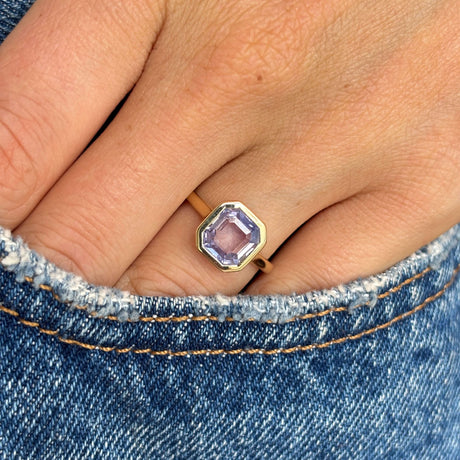 Antique-inspired yellow gold solitaire ring with a square lavender sapphire, styled on hand against blue denim to showcase the gemstone's soft violet hue.