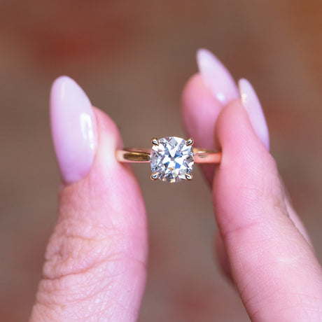 Cushion old-mine cut diamond solitaire engagement ring with square cushion diamond held between fingers.