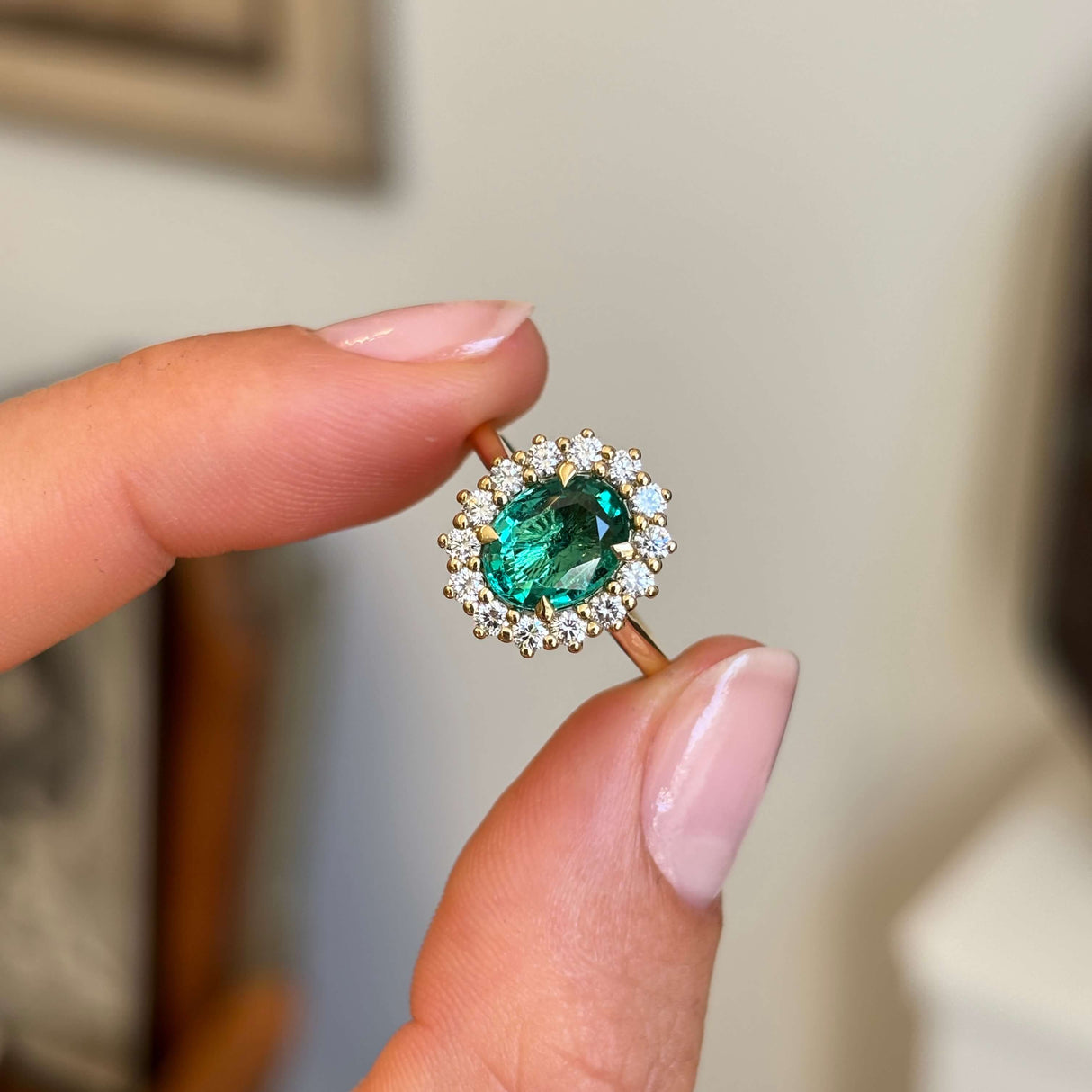 Antique-inspired oval-cut emerald surrounded by brilliant diamonds in a yellow gold cluster setting, held between fingers against a softly blurred vintage interior background.