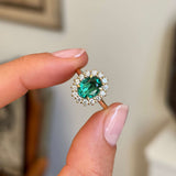 Antique-inspired oval-cut emerald surrounded by brilliant diamonds in a yellow gold cluster setting, held between fingers against a softly blurred vintage interior background.