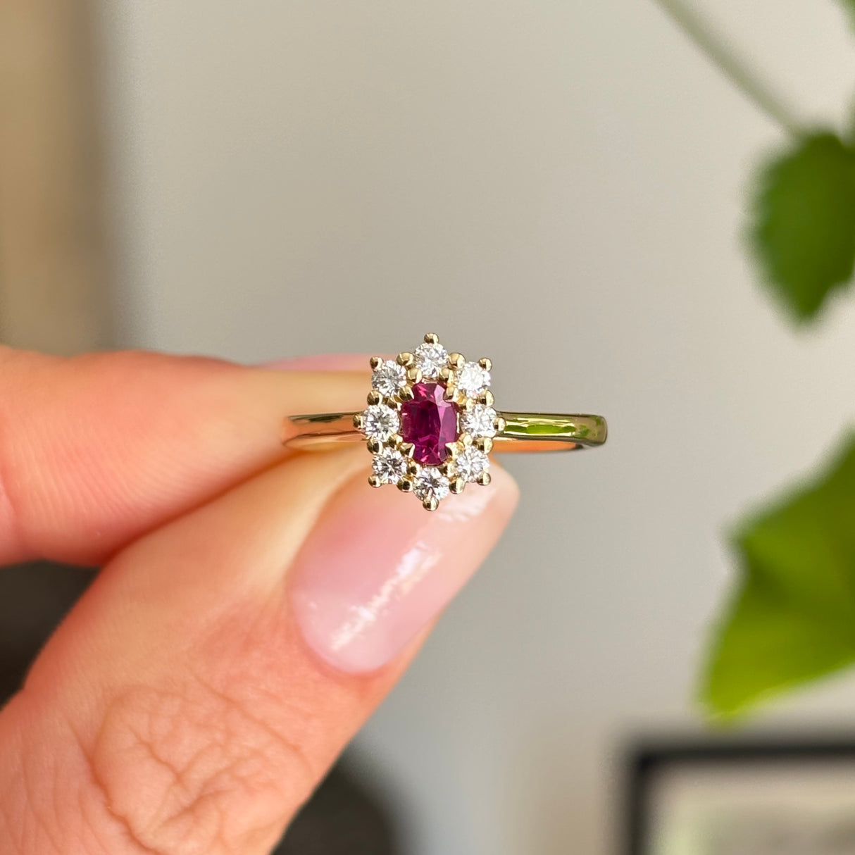 Antique-inspired oval-cut ruby surrounded by brilliant diamonds in a yellow gold cluster setting, held between fingers against a softly blurred interior background with greenery.